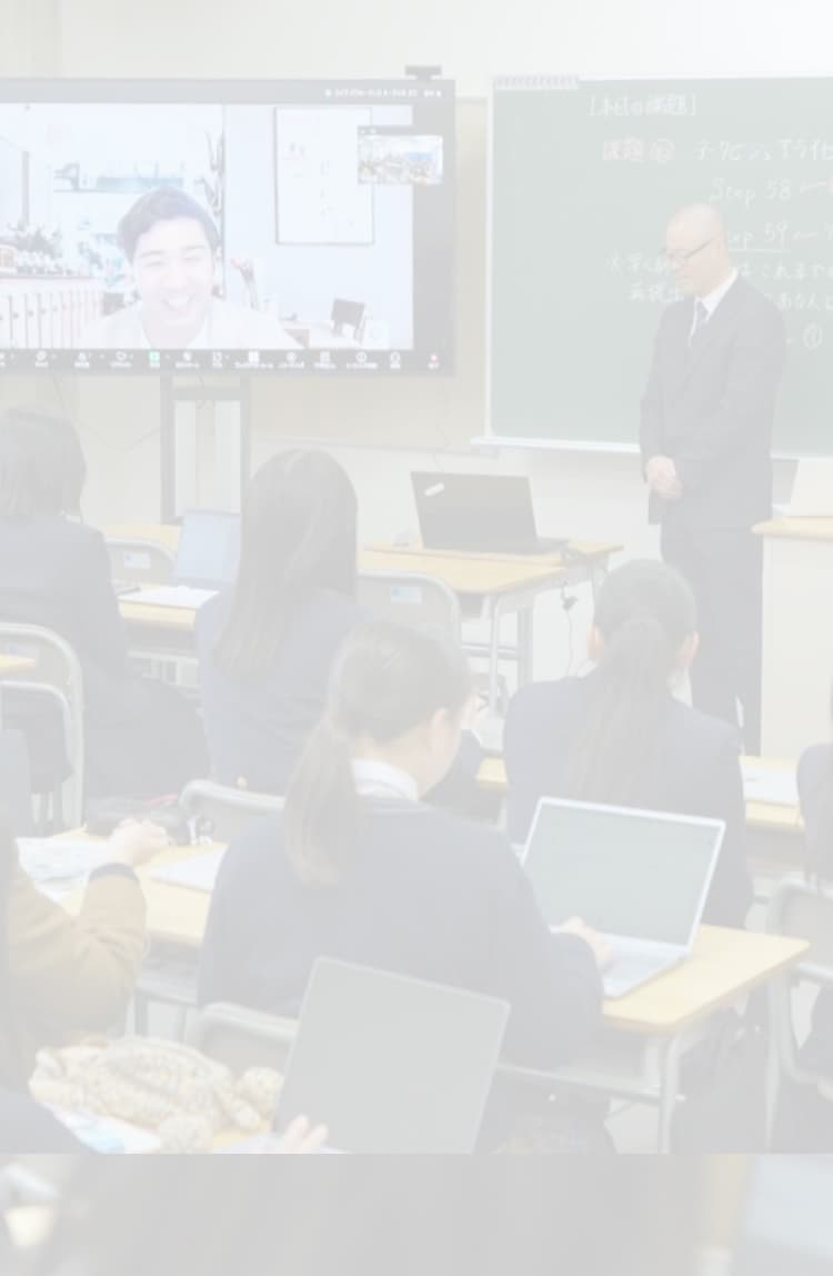 教室で先生が授業を進め、生徒がノートパソコンで学習する様子。大きなディスプレイには学習動画が表示されている。